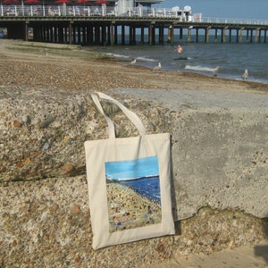 Może przedstawiać: Biała torba z pł&oacute;tna z kolorowym nadrukiem przedstawiającym scenę plażową. Obraz przedstawia pomost, ludzi na plaży i ocean.