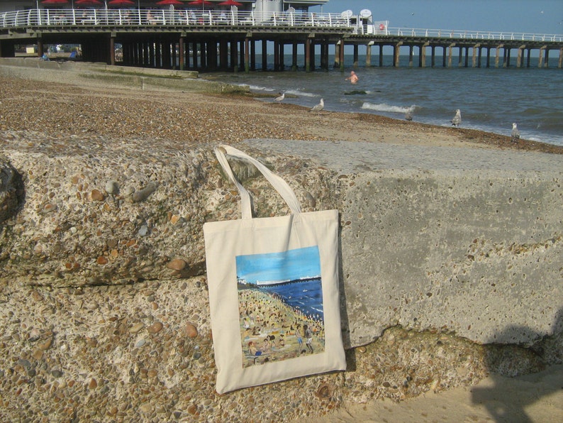 Może przedstawiać: Biała torba z pł&oacute;tna z kolorowym nadrukiem przedstawiającym scenę plażową. Obraz przedstawia pomost, ludzi na plaży i ocean.