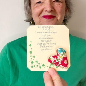 Może przedstawiać: Kremowa karta z napisem "I'm sending you A HUG" i materiałowym sercem z czerwonymi i niebieskimi kwiatowymi wzorami. Karta ma również zielony wzór koniczyny.