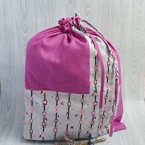 Op de afbeelding: Een grote, roze stoffen opbergtas met een trekkoordsluiting. De tas heeft een paneel met een herhalend patroon van vogelhuisjes op takken. De tas heeft een bijpassend roze trekkoord en accenten.