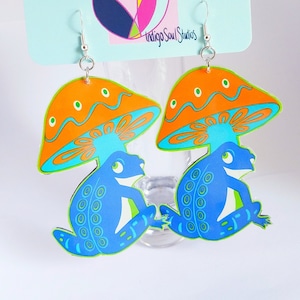 Pode incluir: Um par de brincos extravagantes com um design de cogumelo e rã. Os brincos têm chapéus de cogumelo laranja com detalhes azuis e verdes e corpos de rã azuis. Eles penduram de ganchos prateados.