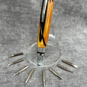 May include: A gold and black pen with a silver top and a set of eight screwdriver bits. The pen is on a clear stand.