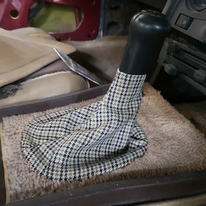 May include: A close-up of a car's gear shift with a black lever and a brown and white checkered gear shift boot.