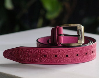 Puede incluir: Un cinturón de cuero rosa con un intrincado diseño floral y una hebilla dorada.