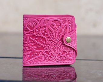 May include: A vibrant pink wallet with an intricate floral pattern embossed on the front. The wallet features a snap closure with a gold-colored button. The design is reflected on a dark, glossy surface.