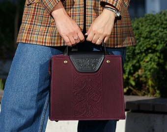 May include: A woman wearing a plaid blazer, blue jeans, and white sneakers is holding a burgundy handbag with a black leather handle and decorative details.
