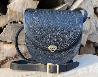 May include: A black leather crossbody bag with intricate floral and geometric embossed details. The bag has a flap closure with a gold metal buckle and a long adjustable strap.