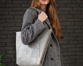 May include: A woman in a gray coat carries a silver tote bag with an embossed floral design.