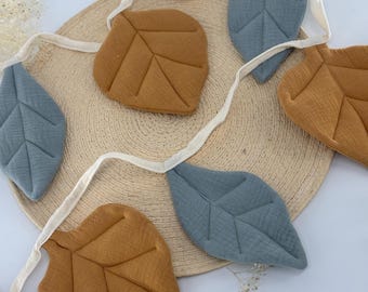 May include: A decorative leaf garland featuring alternating leaves in muted blue and golden brown. The leaves are made of textured fabric and are strung together with a cream-colored ribbon. The garland is arranged on a woven, circular mat.