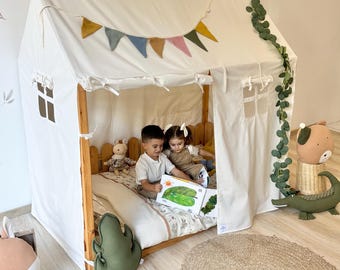 May include: A white playhouse tent with a wooden frame, decorated with a colorful pennant banner and faux greenery. Inside, two children are reading a book. Decorative pillows and stuffed animals add to the cozy atmosphere.