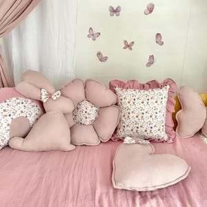 May include: A set of pink and white decorative pillows with floral patterns. The pillows are shaped like a rainbow, a bunny, a flower, a butterfly, and a heart. The pillows are arranged on a pink bedspread.