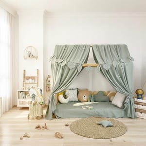 May include: A light green canopy bed with leaf accents and matching bedding is the focal point of a child's room. Decorative pillows, a round jute rug, and wooden toys complete the cozy space. A small teepee and storage baskets are also visible.