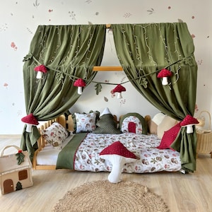 Pode incluir: Uma cama com dossel verde com roupa de cama com padrão de cogumelos brancos e marrons. A cama é decorada com almofadas de cogumelos vermelhos e uma corda de luzes com decorações de cogumelos vermelhos e folhas.