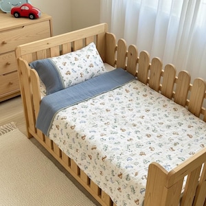 Op de afbeelding: Een houten babybedje met een blauw-witte beddengoedset met auto's. De set bevat een kussen en een deken. Het bedje heeft een hekwerkontwerp. Een rode auto en een wolkvormig nachtlampje staan op een houten commode.