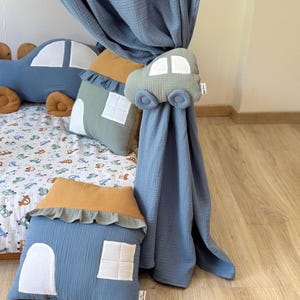 May include: A blue car-shaped pillow and house-shaped pillows with white window details are displayed on a bed with a car-themed patterned sheet. A blue curtain is held back by a car-shaped tieback. The pillows have a ruffled trim.