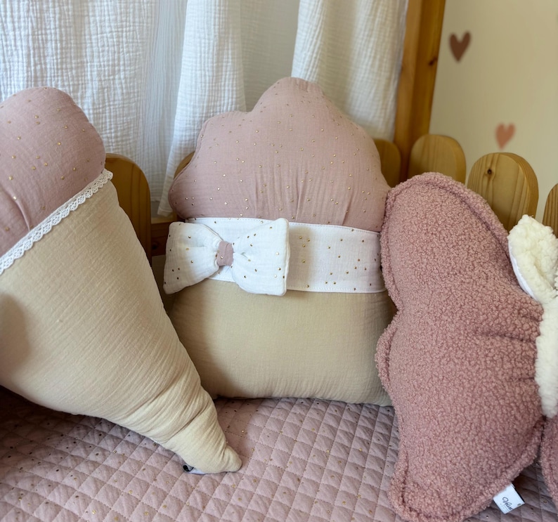 K&ouml;nnte beinhalten: Drei dekorative Kissen in einem rosa und beigefarbenen Farbschema. Die Kissen sind in Form eines Herzens, einer Wolke und eines Sterns. Das Wolkenkissen hat eine wei&szlig;e Schleife und ein wei&szlig;-rosa gepunktetes Muster. Das Sternkissen hat eine flauschige Textur.