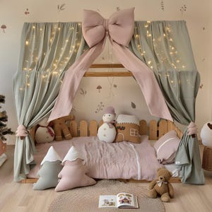 May include: A wooden bed frame with a canopy draped in sage green fabric and a large pink bow. The bed is decorated with plush pillows, a teddy bear, and decorative plush toys. Fairy lights are strung across the canopy.