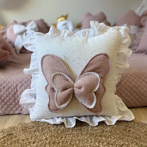 K&ouml;nnte beinhalten: Ein wei&szlig;es Kissen mit einem rosa Schmetterlingsapplikation und goldenen Glitzerakzenten. Das Kissen hat einen wei&szlig;en R&uuml;schenbesatz.