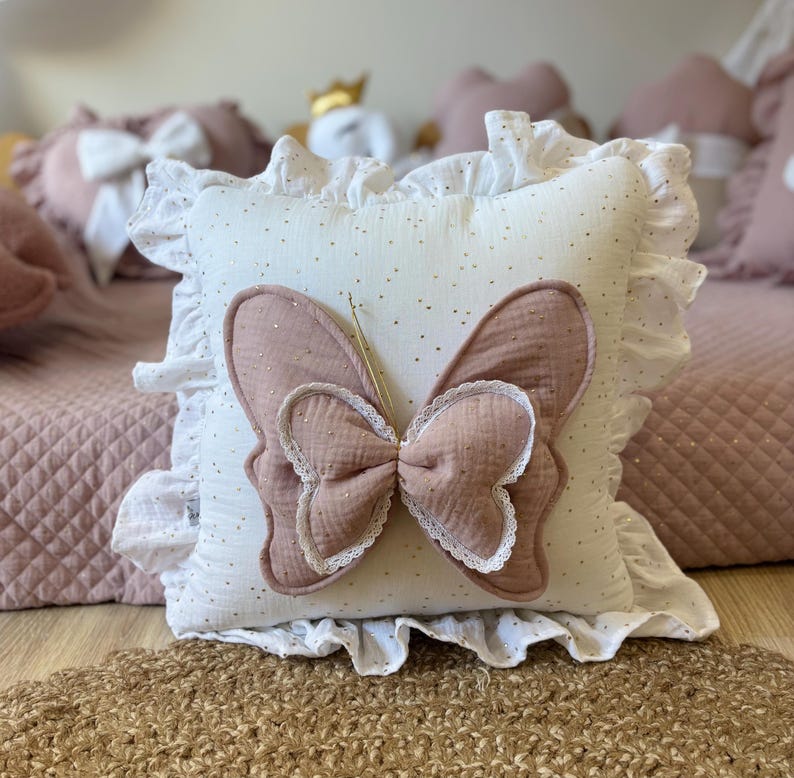 K&ouml;nnte beinhalten: Ein wei&szlig;es Kissen mit einem rosa Schmetterlingsapplikation und goldenen Glitzerakzenten. Das Kissen hat einen wei&szlig;en R&uuml;schenbesatz.