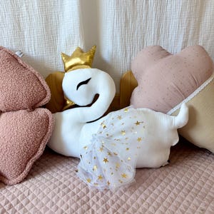 K&ouml;nnte beinhalten: Ein wei&szlig;es Pl&uuml;schschwan-Spielzeug mit einer goldenen Krone und einem wei&szlig;en T&uuml;llrock mit goldenen Sternen. Der Schwan sitzt auf einem rosa gesteppten Bett mit einem rosa-beigen herzf&ouml;rmigen Kissen.