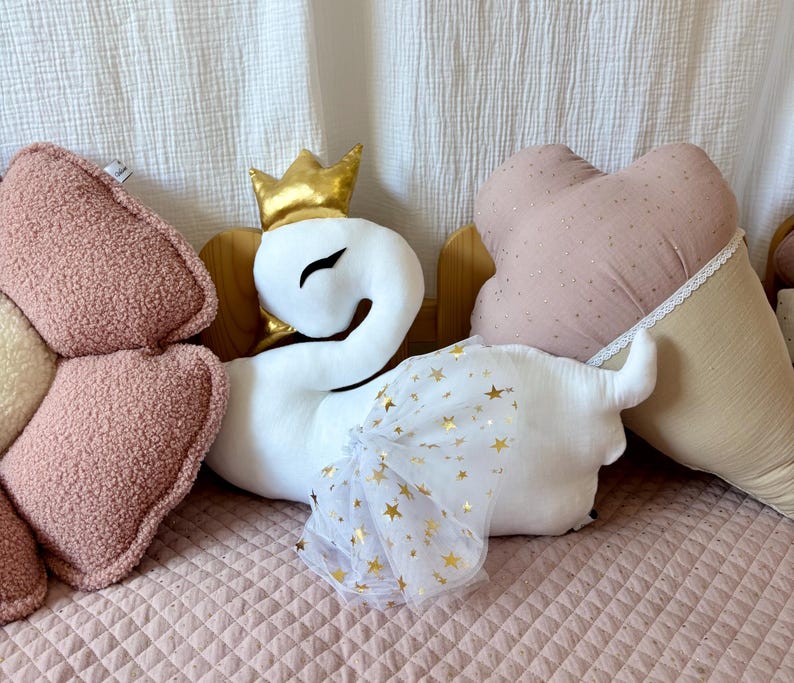 K&ouml;nnte beinhalten: Ein wei&szlig;es Pl&uuml;schschwan-Spielzeug mit einer goldenen Krone und einem wei&szlig;en T&uuml;llrock mit goldenen Sternen. Der Schwan sitzt auf einem rosa gesteppten Bett mit einem rosa-beigen herzf&ouml;rmigen Kissen.