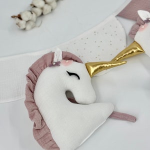 May include: A white unicorn head with a pink ruffled mane and a gold horn. The unicorn has a pink flower and a pearl on its head. The unicorn is part of a garland.