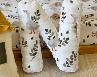 May include: A white, plush letter "N" pillow with a goose and leaf pattern. The pillow is part of a nursery decor set, with a matching patterned background. The goose is gray with orange feet and beak. Green leaves and sprigs are scattered around the goose.