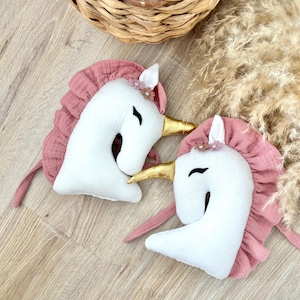 May include: Two white unicorn head plush toys with pink manes and golden horns. Each unicorn has a small floral crown and a curved black eye. The plush toys are on a light wood surface.