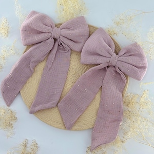 May include: Two mauve-colored hair bows with long, flowing tails, adorned with small, gold-colored embellishments. The bows are made of a textured fabric and are arranged on a round, woven surface, suggesting a craft or accessory theme.