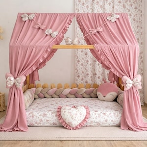 Può includere: Un letto a baldacchino rosa con un materasso a fantasia floreale e un cuscino a forma di cuore. La struttura del letto è in legno e il baldacchino è decorato con fiocchi. Sul letto c'è anche un cuscino a forma di volpe.