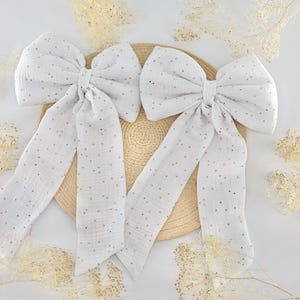 May include: Two large white fabric bows with long streamers, adorned with small, scattered gold-colored dots. The bows are arranged on a round, woven, beige surface, with delicate dried floral accents.