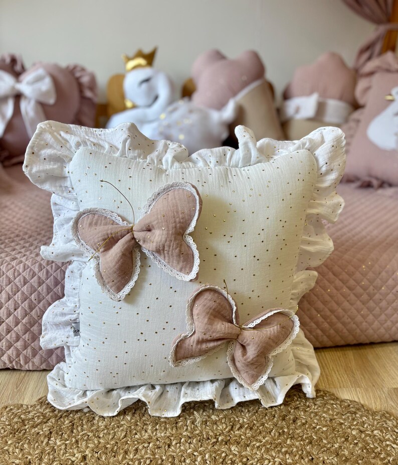 K&ouml;nnte beinhalten: Wei&szlig;es Kissen mit R&uuml;schenrand und zwei rosa Schmetterlingsapplikationen. Das Kissen hat einen wei&szlig;en Hintergrund mit kleinen goldenen Punkten.
