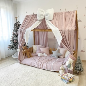 May include: A wooden bed frame with a pink canopy and bedding, decorated with a large white bow. The bed is adorned with pillows, a teddy bear, and a star-shaped pillow. A Christmas tree and decorations are in the background.