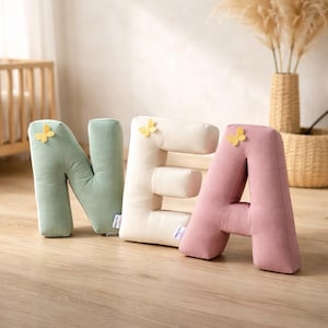 Può includere: Morbide lettere decorative in peluche che compongono la parola "NEA" in colori pastello. Le lettere sono verde chiaro, crema e rosa, ognuna decorata con una piccola farfalla gialla. Sono posizionate su un pavimento in legno, adatte a una cameretta.