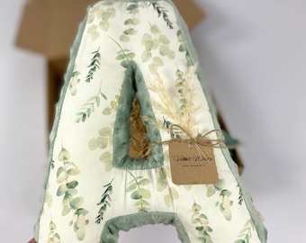 May include: A green and white plush letter A pillow with a leafy pattern. The pillow is surrounded by dried grass and has a small brown tag that says "Velvet White" and a website address.