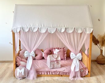 May include: A wooden house-shaped bed frame with a canopy and pink accents. The bed features pink bedding, pillows, and curtains, with white bows and a decorative house-shaped box. The canopy is white with a scalloped edge.