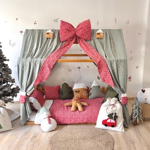 May include: A festive Christmas-themed bed canopy with a wooden frame, draped with sage green fabric and a large red bow. The bed is adorned with decorative pillows, a gingerbread man, and a snowman. A Christmas tree and gift bags complete the holiday scene.
