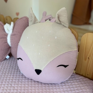 K&ouml;nnte beinhalten: Ein pl&uuml;schiges rosa und beiges Kissen in Form eines Rehs mit gestickten Augen und einer schwarzen Nase. Das Kissen ist rund und hat einen weichen, strukturierten Stoff.