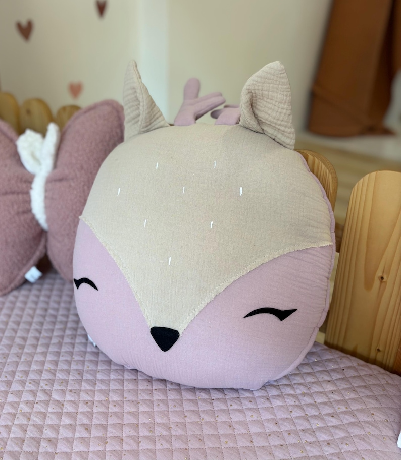 K&ouml;nnte beinhalten: Ein pl&uuml;schiges rosa und beiges Kissen in Form eines Rehs mit gestickten Augen und einer schwarzen Nase. Das Kissen ist rund und hat einen weichen, strukturierten Stoff.