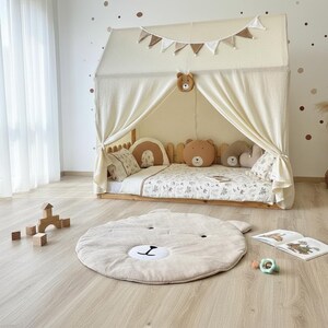 Puede incluir: Una cama infantil con estructura en forma de casa, cubierta con tela color crema. La cama est&aacute; decorada con cojines con forma de oso y una alfombra a juego. Un juguete de madera y un libro est&aacute;n en el suelo.