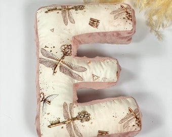 May include: A pink plush letter E pillow with a white fabric front featuring a pattern of dragonfly and key designs.