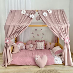 Puede incluir: Una cama con dosel rosa con decoraciones florales y de mariposas. La cama presenta un marco de madera, tela rosa drapeada y almohadas decorativas en varias formas, incluyendo flores, mariposas y un corazón. Una guirnalda de flores blancas y rosas cuelga.