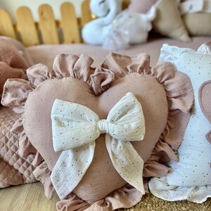 K&ouml;nnte beinhalten: Ein rosa herzf&ouml;rmiges Kissen mit einer wei&szlig;en Schleife und goldenen Glitzerakzenten. Das Kissen hat einen gekr&auml;uselten Rand und ist aus einem weichen, strukturierten Stoff gefertigt.