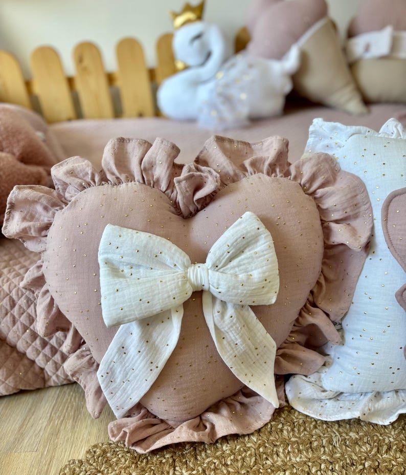 K&ouml;nnte beinhalten: Ein rosa herzf&ouml;rmiges Kissen mit einer wei&szlig;en Schleife und goldenen Glitzerakzenten. Das Kissen hat einen gekr&auml;uselten Rand und ist aus einem weichen, strukturierten Stoff gefertigt.