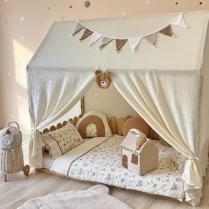 May include: A beige house-shaped bed frame with a canopy, draped with cream-colored fabric. The bed is decorated with bear-themed pillows, a blanket, and a house-shaped toy. A wicker basket with a bear face is on the left.