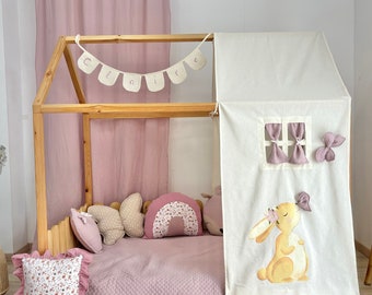 May include: A wooden house-shaped bed frame with a white fabric roof and side panel featuring a bunny illustration. The bed is decorated with pink bedding, pillows, and a banner that spells "Claire".