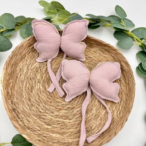 Puede incluir: Dos adornos de mariposa de tela rosa con lazos largos de tela. Las mariposas están sobre una superficie de paja tejida.