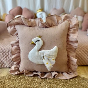 K&ouml;nnte beinhalten: Ein rosa Kissen mit einer wei&szlig;en Schwanenapplikation und goldenen Glitzerakzenten. Das Kissen hat einen R&uuml;schenrand und ist mit goldenen Sternen verziert.