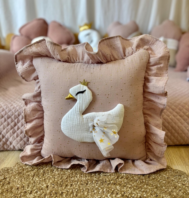 K&ouml;nnte beinhalten: Ein rosa Kissen mit einer wei&szlig;en Schwanenapplikation und goldenen Glitzerakzenten. Das Kissen hat einen R&uuml;schenrand und ist mit goldenen Sternen verziert.