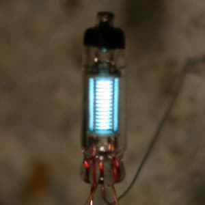 Peut inclure: Un tube à vide lumineux avec un dessus noir et un corps en verre transparent. Les composants internes émettent une lumière bleu vif. De fins fils s'étendent de la base.